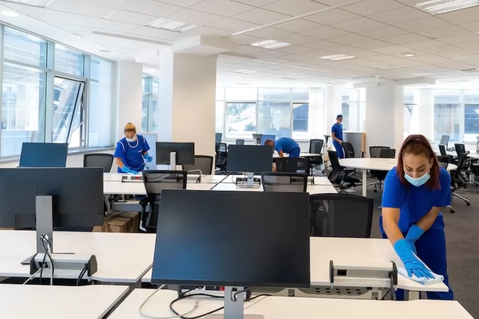 Professional cleaners wearing medical masks and gloves cleaning desks in large empty office.
